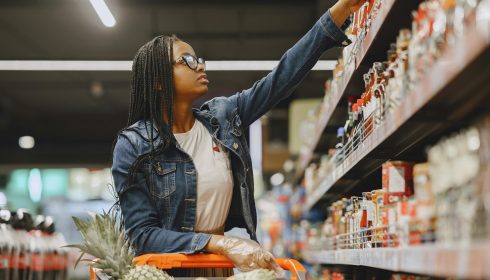 A woman wearing glasses and a denim jacket reaches for an item on a supermarket shelf while pushing a shopping trolley filled with groceries.