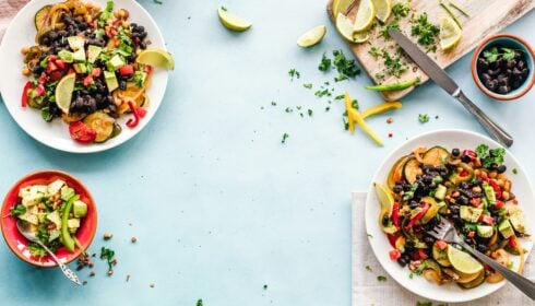 Three plates of colourful salad with beans, vegetables, avocado, and lime wedges on a blue surface, with a chopping board, knife, and scattered herbs nearby.