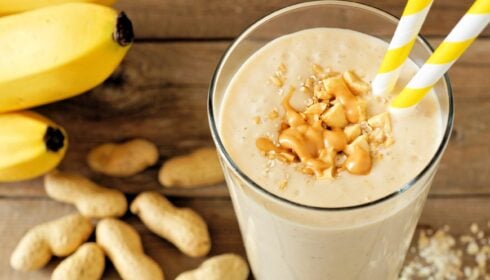 A glass of peanut butter banana smoothie with yellow-striped straws, topped with peanuts and granola, sits on a wooden surface with bananas and peanuts nearby.