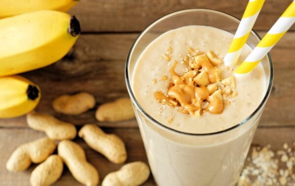 A glass of peanut butter banana smoothie with yellow-striped straws, topped with peanuts and granola, sits on a wooden surface with bananas and peanuts nearby.