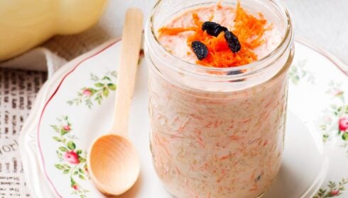 A glass jar of overnight oats topped with grated carrot and sultanas sits on a floral plate with a wooden spoon beside it.