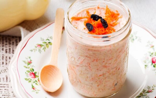 A glass jar of overnight oats topped with grated carrot and sultanas sits on a floral plate with a wooden spoon beside it.