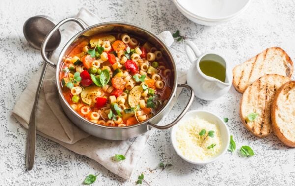 A pot of vegetable and pasta soup with herbs, served with grilled bread, grated cheese, and a small jug of olive oil on a textured white surface.