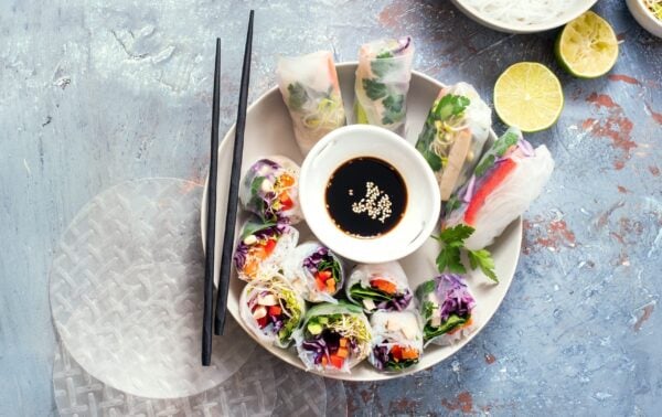 Plate of vegetable spring rolls with soy dipping sauce in the centre, black chopsticks on the side, and sliced limes and rice paper sheets nearby.