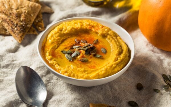 A bowl of pumpkin houmous topped with seeds and paprika, surrounded by pitta chips and a spoon on a linen cloth.