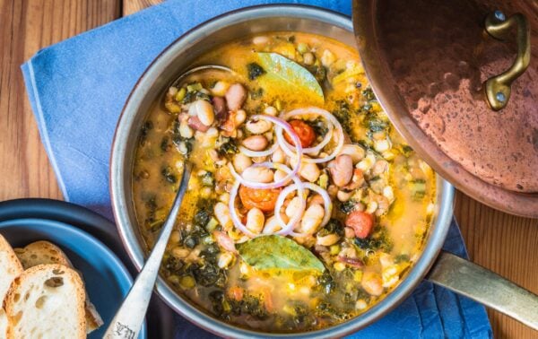 A pot of vegetable and bean stew with bay leaves and red onion slices, served on a blue cloth with toasted bread on the side.