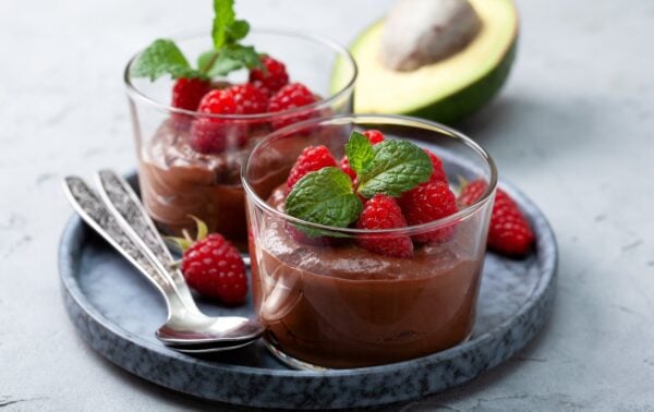 Two glass tumblers of chocolate mousse topped with raspberries and mint leaves on a grey plate with two spoons, fresh raspberries, and half an avocado in the background.