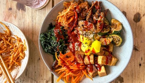 A bowl with tofu, sliced carrots, courgette, mushrooms, greens, pickled radish, noodles, and sauce, topped with sesame seeds. A side plate of noodles and drink are visible on a wooden table.