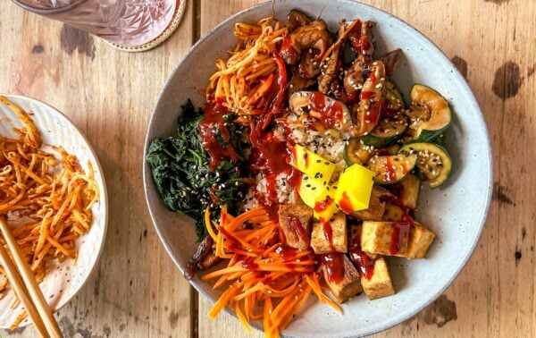 A bowl with tofu, sliced carrots, courgette, mushrooms, greens, pickled radish, noodles, and sauce, topped with sesame seeds. A side plate of noodles and drink are visible on a wooden table.