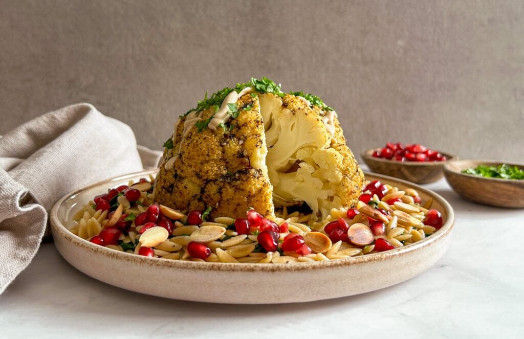 Roasted whole cauliflower served on orzo pasta, garnished with pomegranate seeds, almonds, and chopped herbs. A bowl of pomegranate seeds and herbs are in the background.