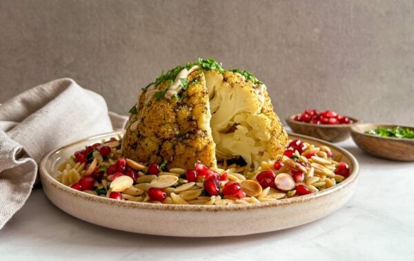 Roasted whole cauliflower served on orzo pasta, garnished with pomegranate seeds, almonds, and chopped herbs. A bowl of pomegranate seeds and herbs are in the background.