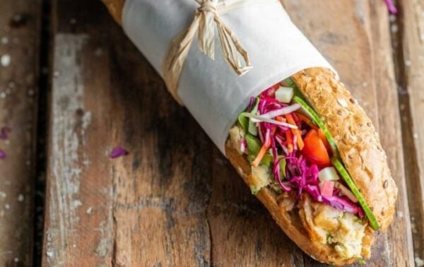 Wholegrain sandwich filled with shredded chicken, spinach, red cabbage, cucumber, and carrots, wrapped in greaseproof paper and tied with string, on a rustic wooden board.