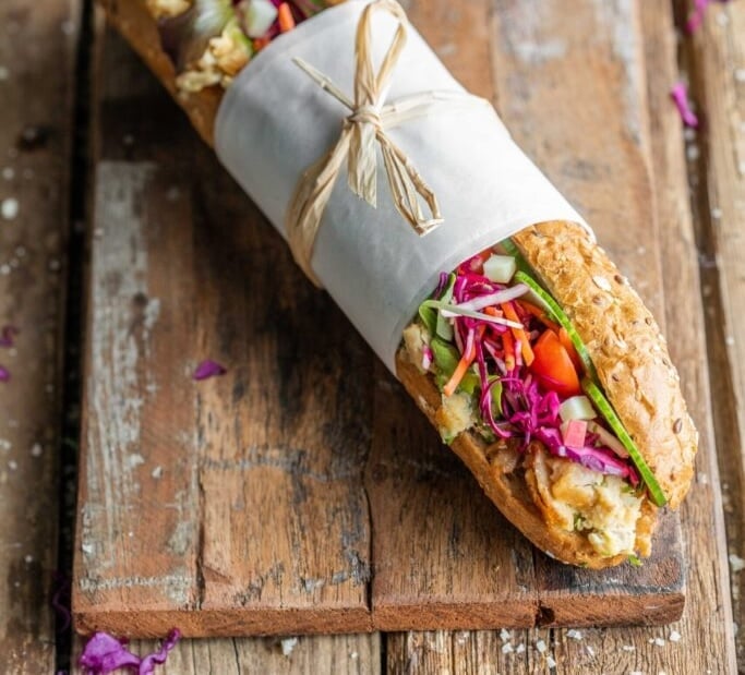 Wholegrain sandwich filled with shredded chicken, spinach, red cabbage, cucumber, and carrots, wrapped in greaseproof paper and tied with string, on a rustic wooden board.