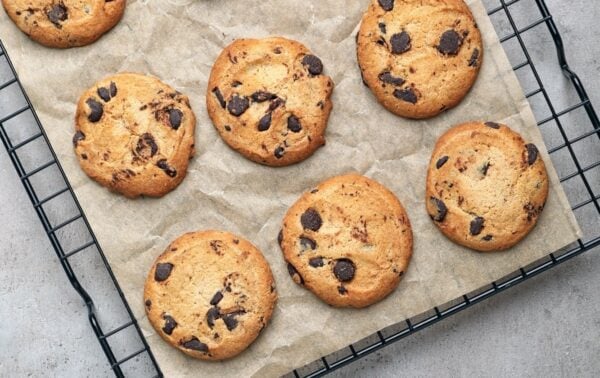 Nine chocolate chip biscuits on baking parchment atop a black wire cooling rack, viewed from above on a grey surface.
