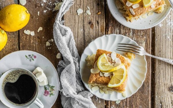 Slices of lemon cake with almond flakes on plates, cups of black coffee, lemon halves, and a grey serviette on a rustic wooden table.