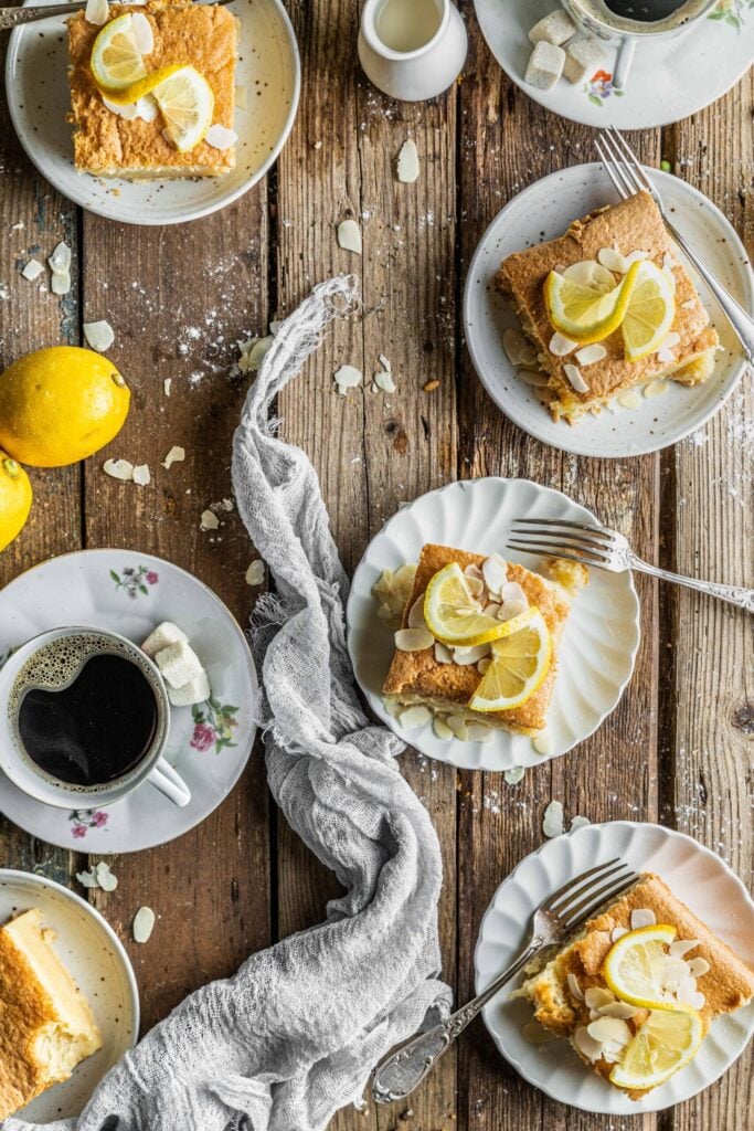 Slices of lemon cake with almond flakes on plates, cups of black coffee, lemon halves, and a grey serviette on a rustic wooden table.