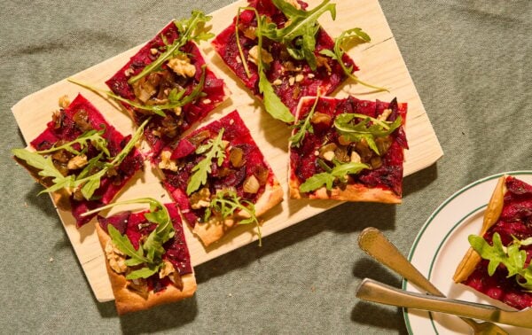 Rectangular flatbread slices with beet topping, walnuts, caramelised onions, and rocket are arranged on a wooden board, with a few slices on a nearby plate with cutlery.