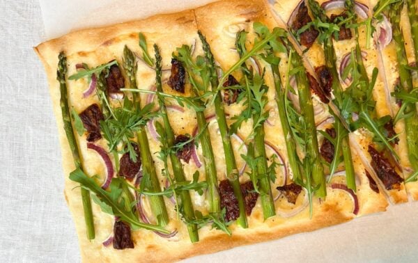 Rectangular flatbread topped with asparagus, rocket, red onion slices, and sun-dried tomatoes, placed on baking paper.