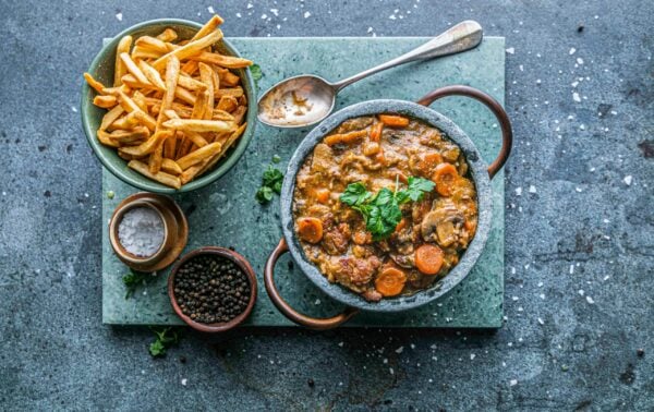 A bowl of beef stew with carrots, garnished with herbs, next to a bowl of chips, a spoon, and small bowls of salt and pepper on a stone board.