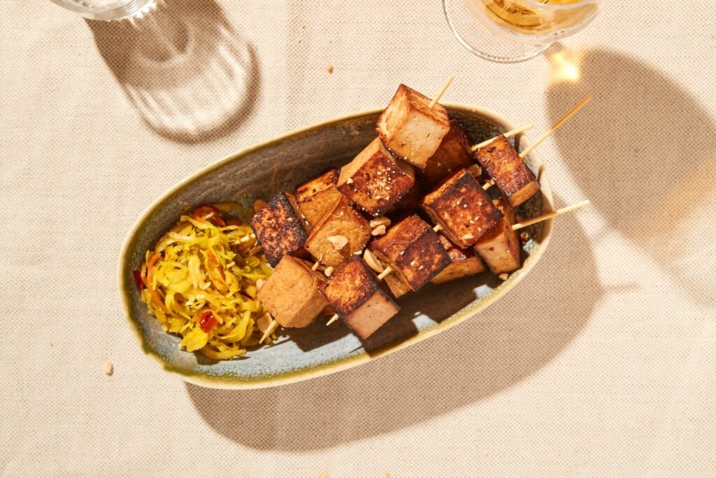 Oval plate with grilled tofu kebabs and a side of shredded vegetable salad, placed on a beige tablecloth under natural light.