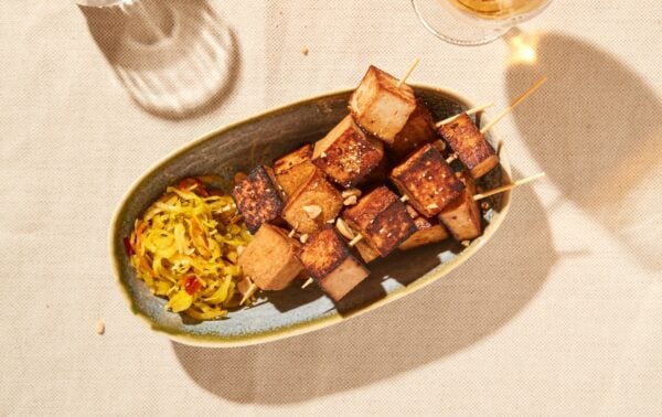 Oval plate with grilled tofu kebabs and a side of shredded vegetable salad, placed on a beige tablecloth under natural light.