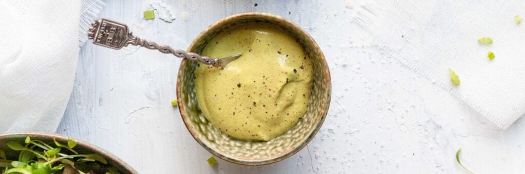 A bowl of creamy green salad dressing with a spoon, topped with ground black pepper, sits on a white textured surface.