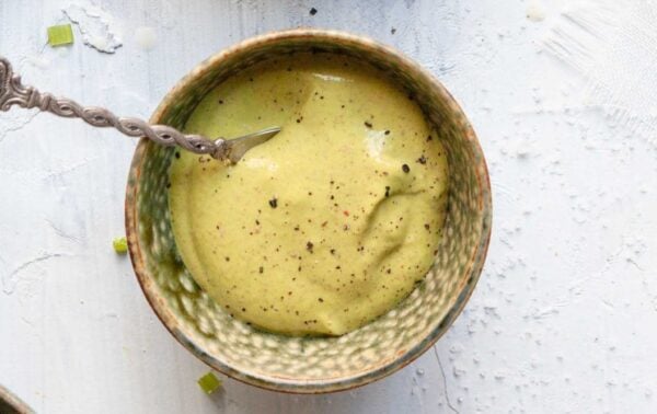 A bowl of creamy green salad dressing with a spoon, topped with ground black pepper, sits on a white textured surface.