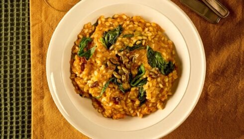 A white bowl of risotto topped with wilted greens and seeds, set on a tan serviette with cutlery and a green textured place mat nearby.
