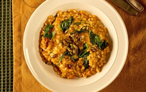 A white bowl of risotto topped with wilted greens and seeds, set on a tan serviette with cutlery and a green textured place mat nearby.