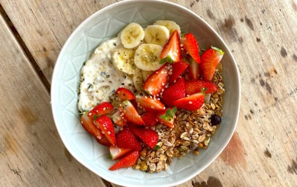 A bowl of yoghurt topped with sliced bananas, strawberries, granola, and seeds sits on a wooden table.