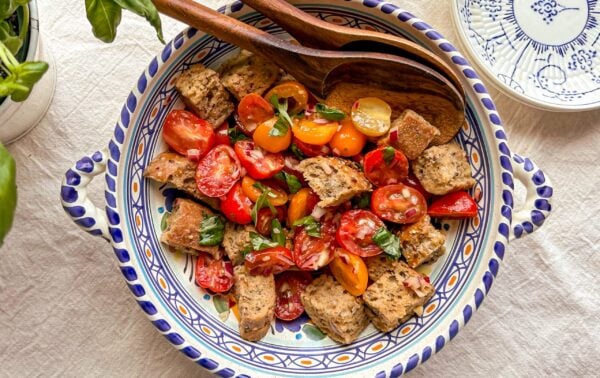 A bowl of panzanella salad with cherry tomatoes, bread cubes, basil, and wooden serving utensils on a patterned plate.