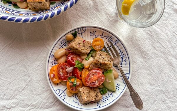 A plate with tomato, cucumber, white beans, herbs, and croutons sits next to a fork; a glass of water with a lemon slice is nearby.