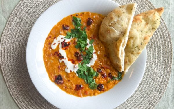 A white bowl of lentil curry garnished with coriander and cream, served with two pieces of naan bread on a woven placemat.