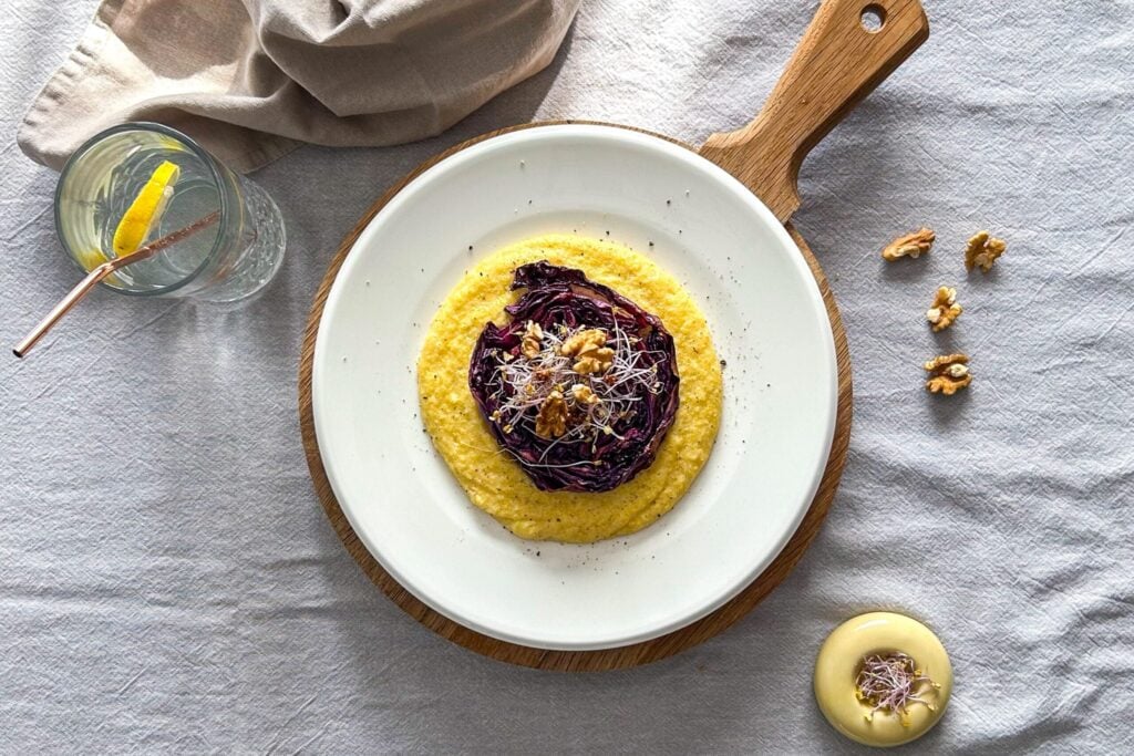 A plate of creamy yellow polenta topped with roasted red cabbage, walnuts, and sprouts, served on a wooden board with a glass of lemon water and extra walnuts on the side.