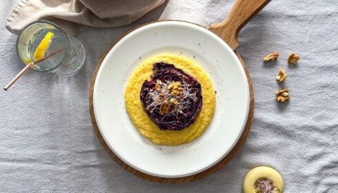 A plate of creamy yellow polenta topped with roasted red cabbage, walnuts, and sprouts, served on a wooden board with a glass of lemon water and extra walnuts on the side.