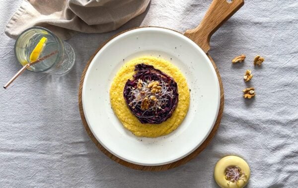 A plate of creamy yellow polenta topped with roasted red cabbage, walnuts, and sprouts, served on a wooden board with a glass of lemon water and extra walnuts on the side.