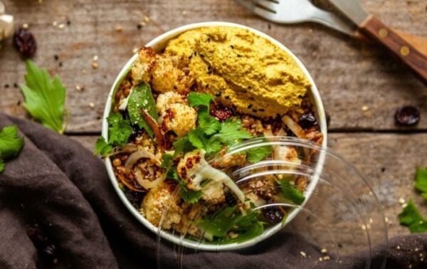 A bowl of roasted cauliflower, grains, greens, seeds, and a scoop of yellow houmous sits on a rustic wooden table with a fork and greens nearby.