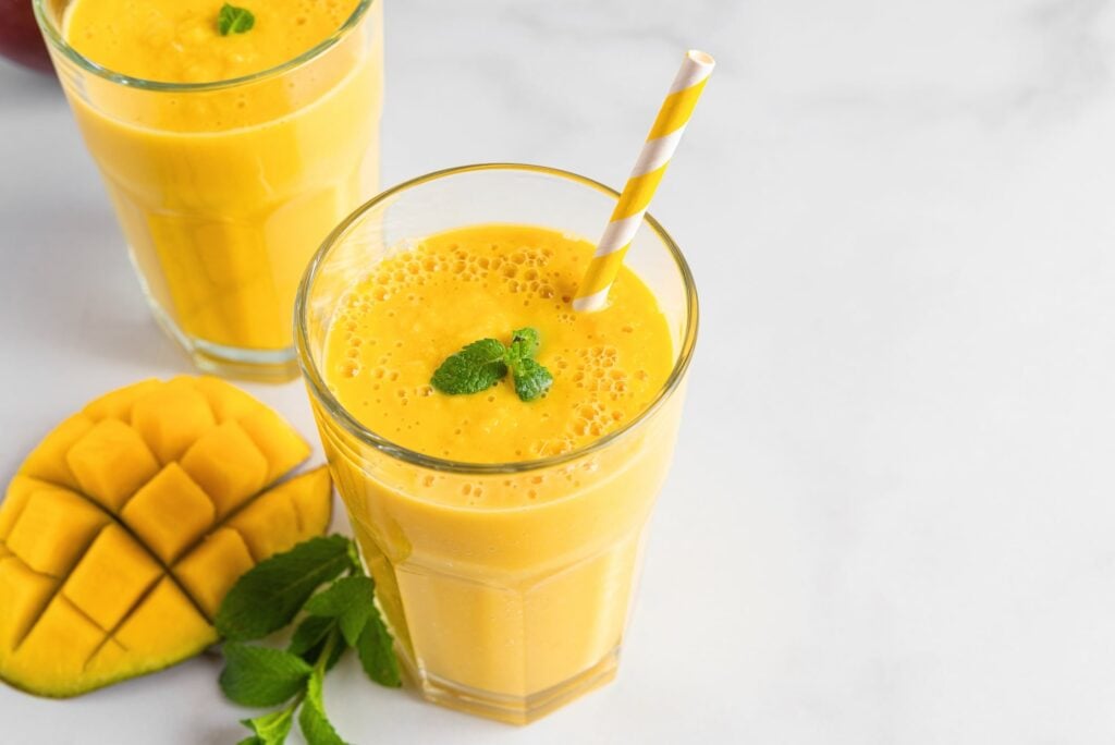 A glass of mango smoothie with a striped straw, garnished with mint, next to a sliced mango and mint leaves on a white surface. Another smoothie is in the background.