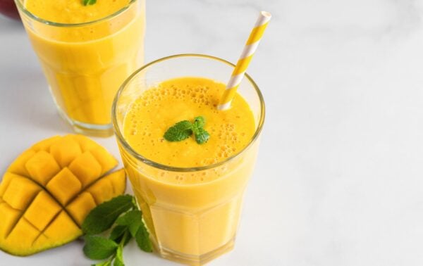 A glass of mango smoothie with a striped straw, garnished with mint, next to a sliced mango and mint leaves on a white surface. Another smoothie is in the background.