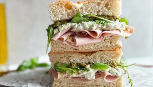 A sandwich with slices of ham, leafy greens, white sauce, and pesto is stacked on focaccia bread, cut in half and placed on greaseproof paper.