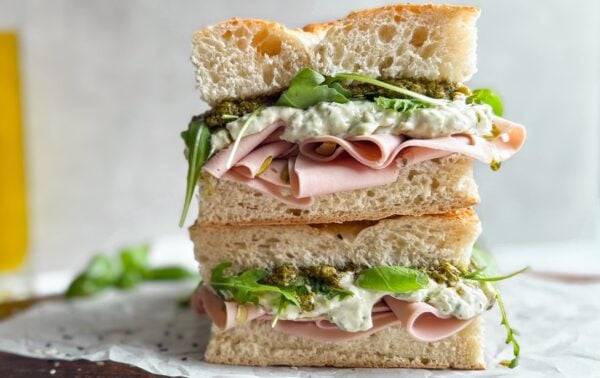 A sandwich with slices of ham, leafy greens, white sauce, and pesto is stacked on focaccia bread, cut in half and placed on greaseproof paper.