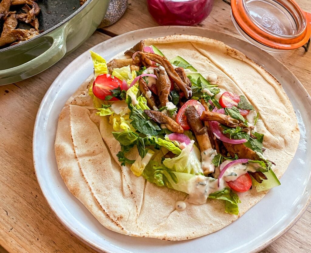 A flatbread topped with lettuce, cherry tomatoes, onions, herbs, grilled mushrooms, and creamy dressing is served on a white plate.