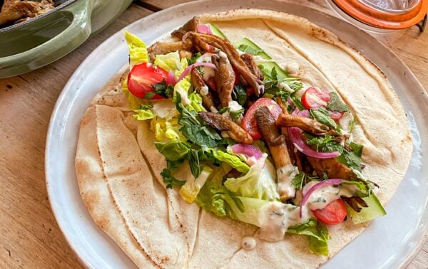 A flatbread topped with lettuce, cherry tomatoes, onions, herbs, grilled mushrooms, and creamy dressing is served on a white plate.