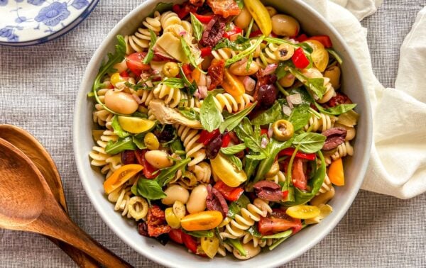 Bowl of fusilli pasta salad with cherry tomatoes, beans, olives, basil, and greens, placed on a grey tablecloth beside wooden utensils and a white serviette.