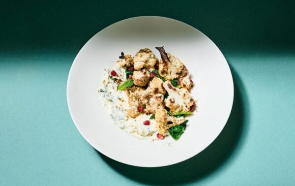 A white plate with roasted cauliflower, greens, pomegranate seeds, and a creamy white sauce, set against a teal background.