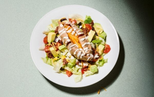 A baked sweet potato with soured cream is served on a salad of lettuce, cherry tomatoes, sweetcorn, avocado, cheese, and black beans, on a white plate.