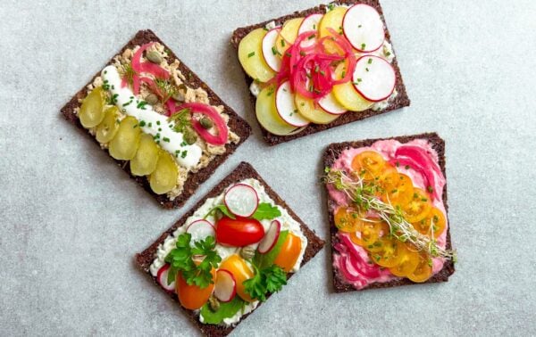 Four open-faced sandwiches on dark bread topped with a variety of sliced vegetables, spreads, and herbs, arranged on a light grey surface.