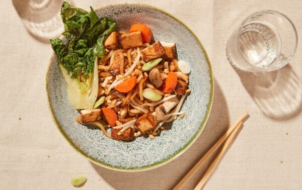 A bowl of stir-fried noodles with tofu, pak choi, carrots, and spring onions, served on a blue plate with a glass of water and chopsticks on a beige tablecloth.