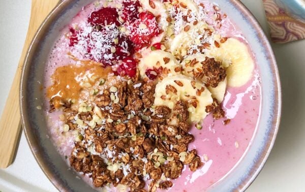 Smoothie bowl topped with banana slices, raspberries, desiccated coconut, granola, peanut butter, and seeds in a ceramic bowl on a light surface.