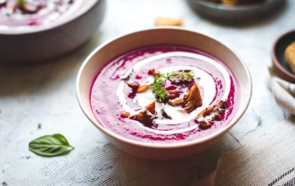 Bowl of purple soup garnished with cream, mushrooms, and herbs on a light table, with a serviette and bread visible in the background.